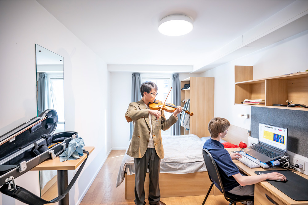 Two male students playing the violin and one recording on their laptop in a bedroom.